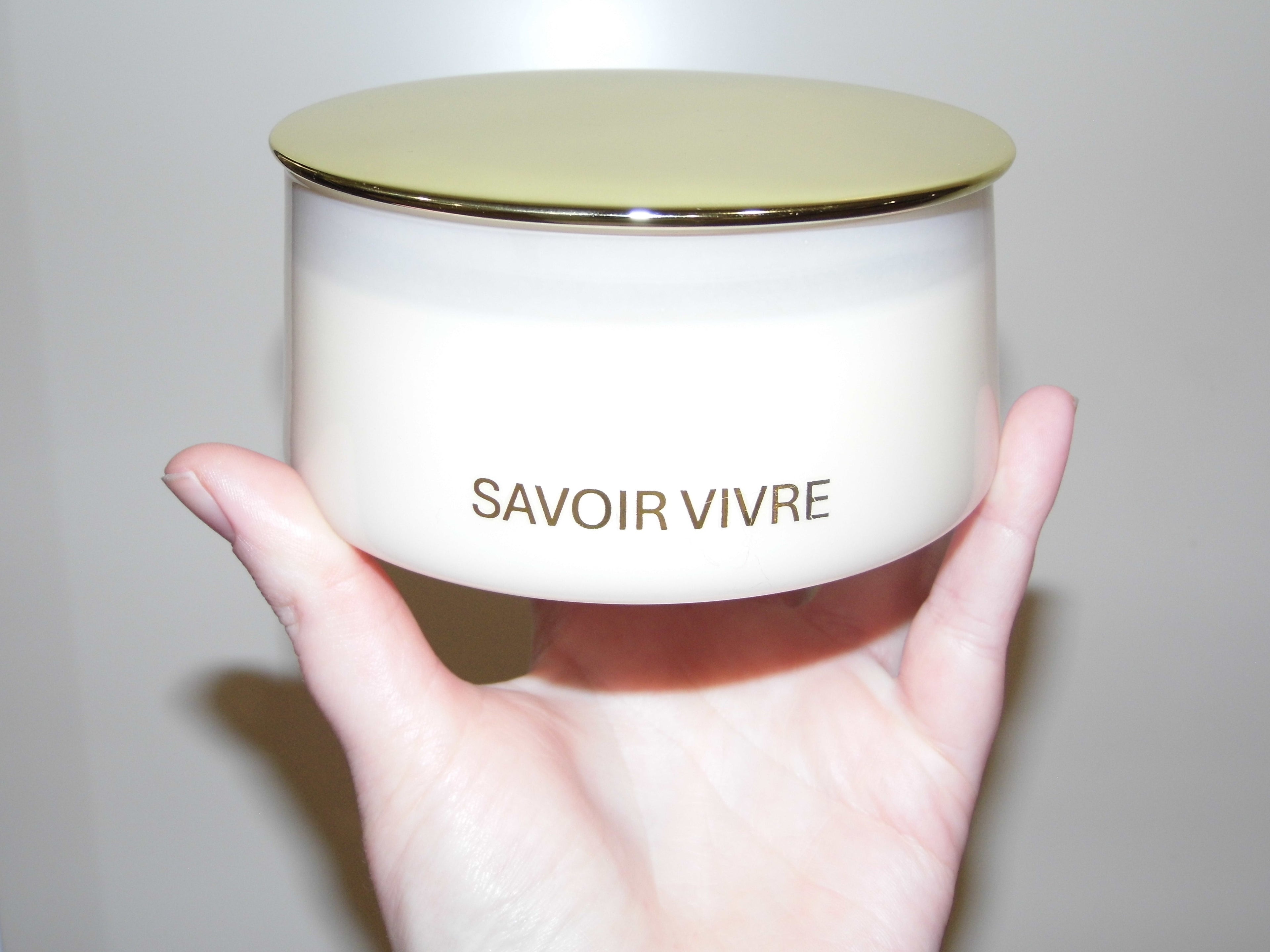 Hand holding a blush NZ candle jar with 'Savoir Vivre' branding against a plain background